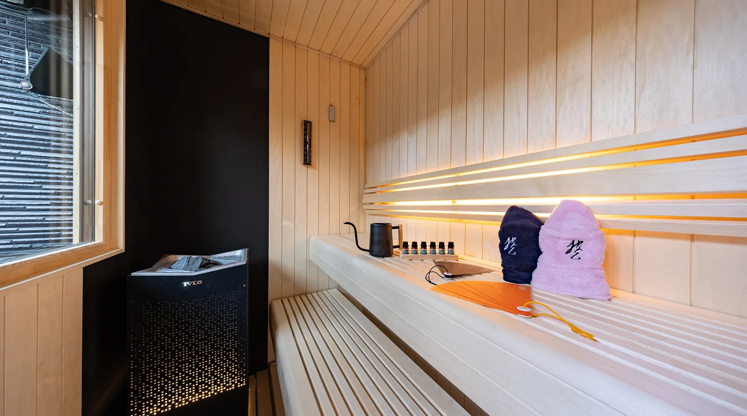間接照明が灯る木造のプライベートサウナ。室内にサウナストーン、ロウリュ用の水差し、サウナハットが備え付けられている様子