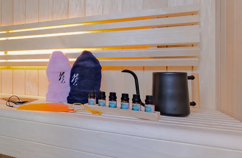 ロウリュ用の黒い水差し、6種類のアロマオイル、ピンクと紺のサウナハット、オレンジ色のうちわが並ぶサウナ室内の備品